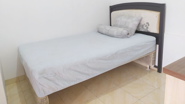 A small, single-bed room with a plain white mattress and a checkered pillow. The bed has a simple wooden headboard and is positioned against a plain wall. - Hotel O Homestay Amanda Syariah (Makassar)