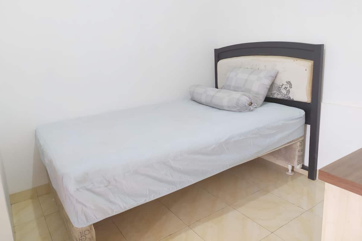 A small, single-bed room with a plain white mattress and a checkered pillow. The bed has a simple wooden headboard and is positioned against a plain wall.