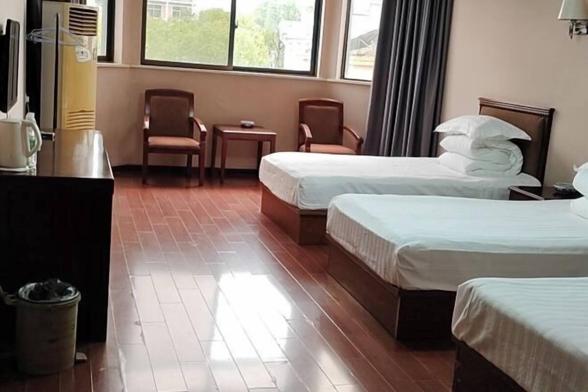 A hotel room with two beds, a desk, chairs, a trash bin, and a window with a view.