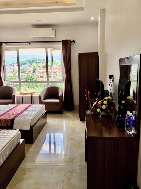 Habitación de hotel con dos camas, un escritorio con computadora y espejo, una silla y un ventanal con vistas a edificios y vegetación.