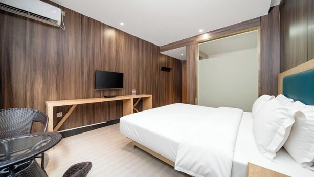 Habitación de hotel moderna con panel de pared de madera, televisor de pantalla plana, cama con sábanas blancas, un pequeño escritorio de madera y una mesa redonda con sillas de mimbre.