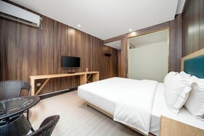 Une chambre d’hôtel moderne avec un panneau mural en bois, un téléviseur à écran plat, un lit avec du linge de maison blanc, un petit bureau en bois et une table ronde avec des chaises en osier.