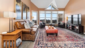A spacious living room with a large window, a leather sofa, a red ottoman, and a TV stand with a flat-screen TV. - Shenandoah Ridge 11 with Wintergreen Advantage (Wintergreen)