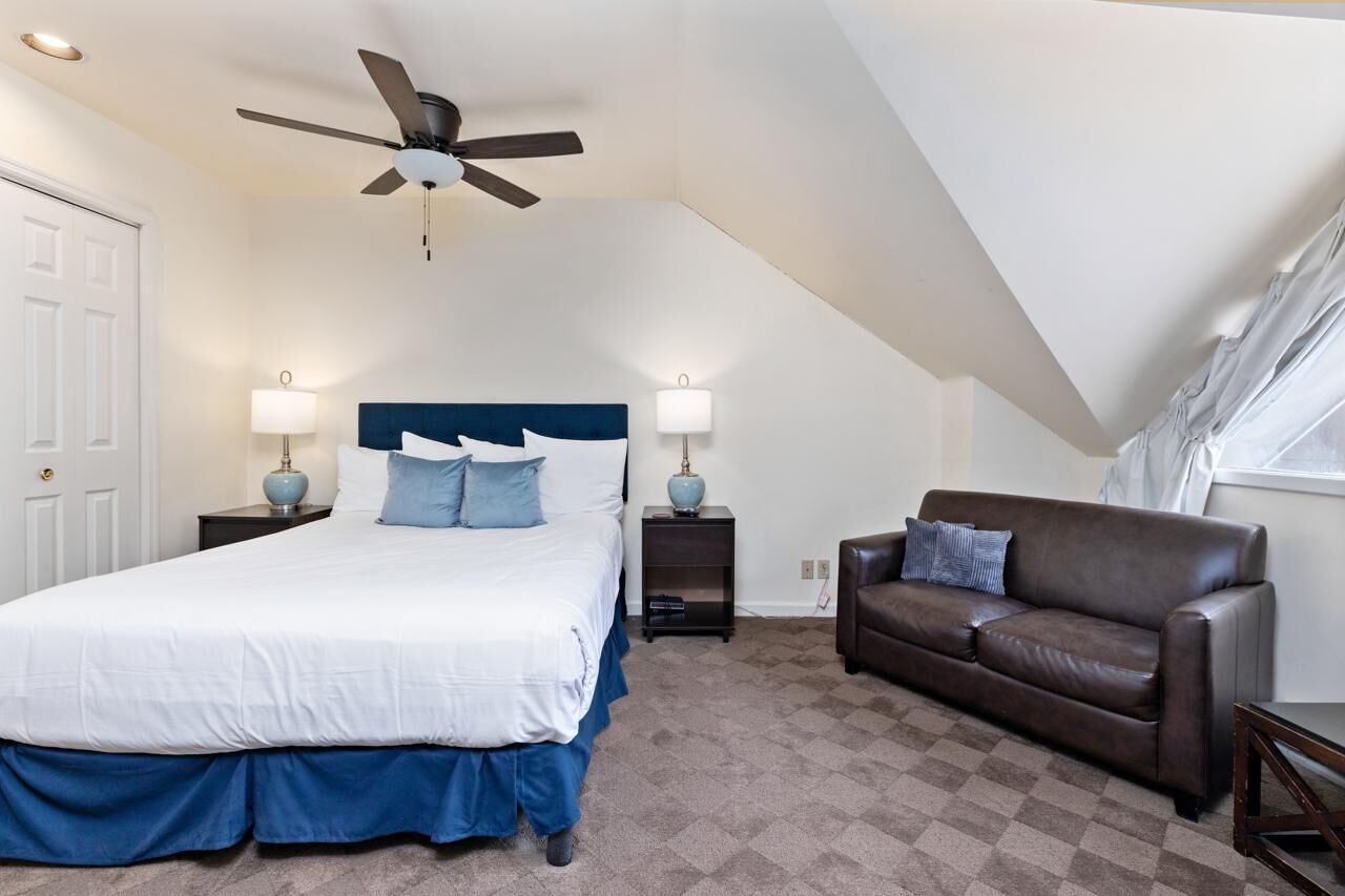 A bedroom with a bed, a ceiling fan, a brown leather couch, and two bedside lamps.