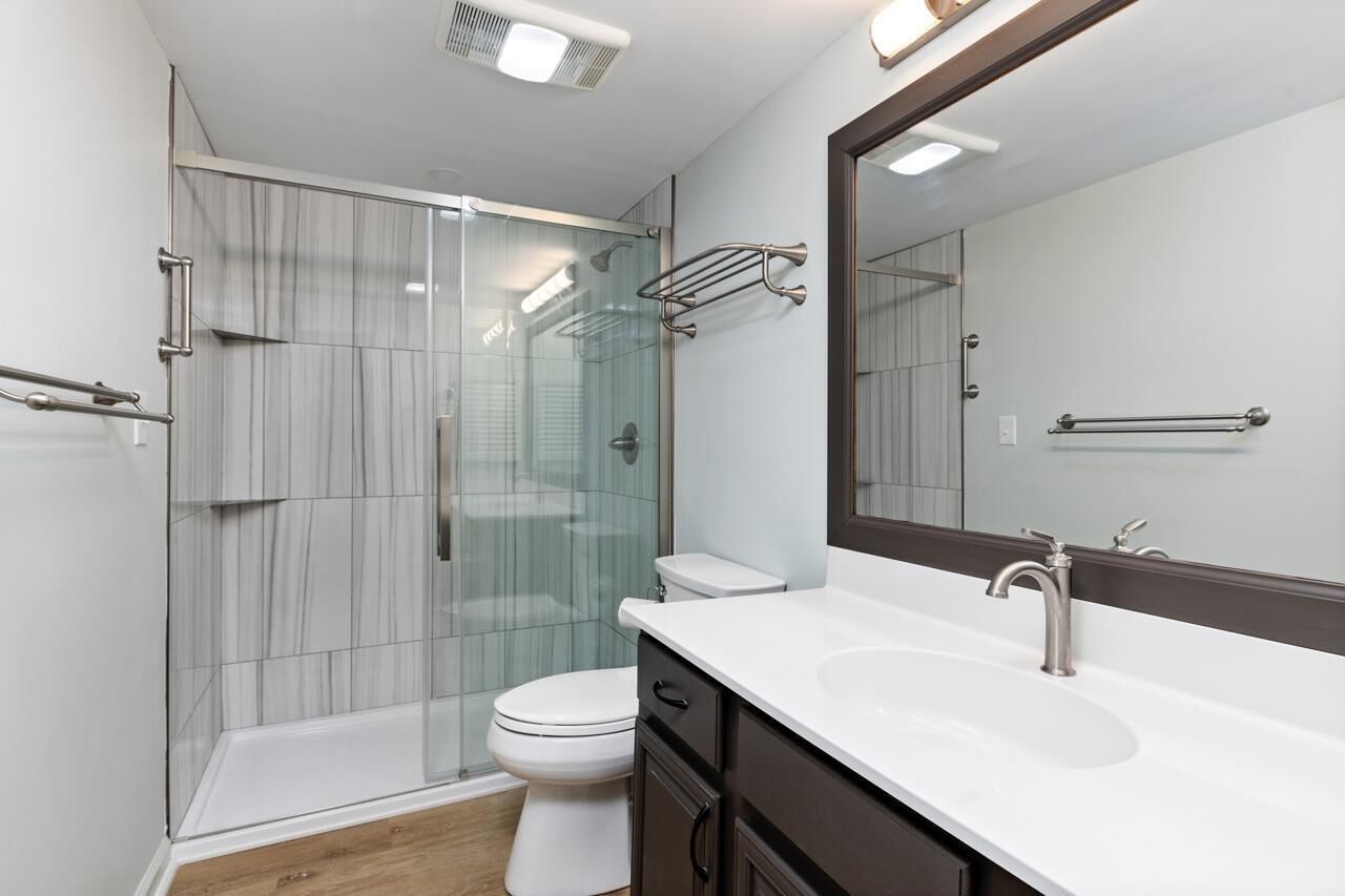 Un baño moderno con cabina de ducha de vidrio, inodoro blanco, tocador con un espejo grande y lavamanos.