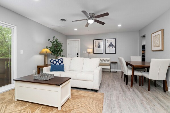 A living room with a white sofa, a coffee table, a dining table with chairs, a ceiling fan, and framed pictures on the wall.