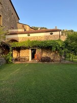 Un edificio rústico de piedra con techo de tejas, un pequeño jardín y mobiliario exterior.