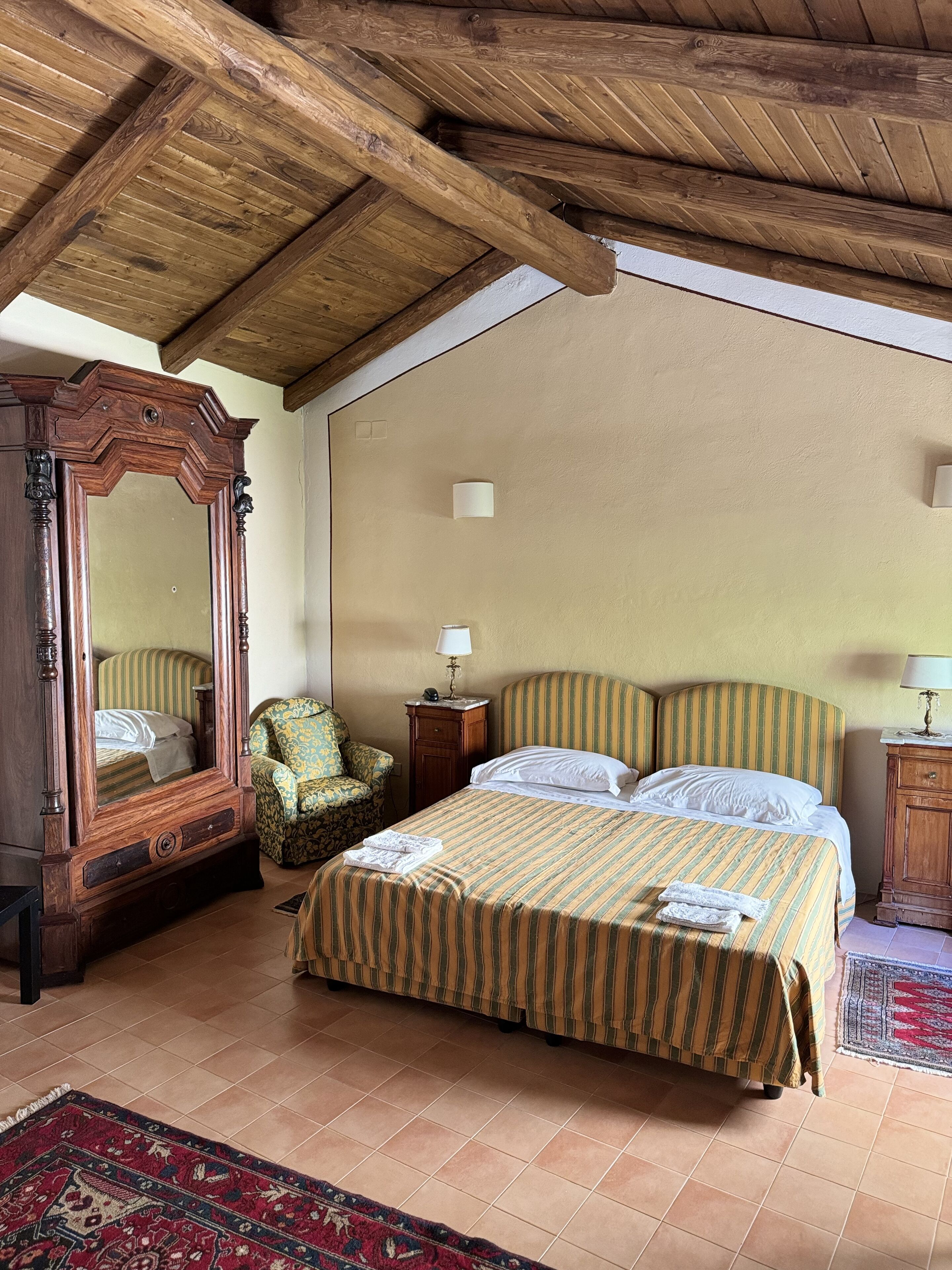 A bedroom with a wooden ceiling, a large mirror, a striped bed, a chair, and a nightstand.