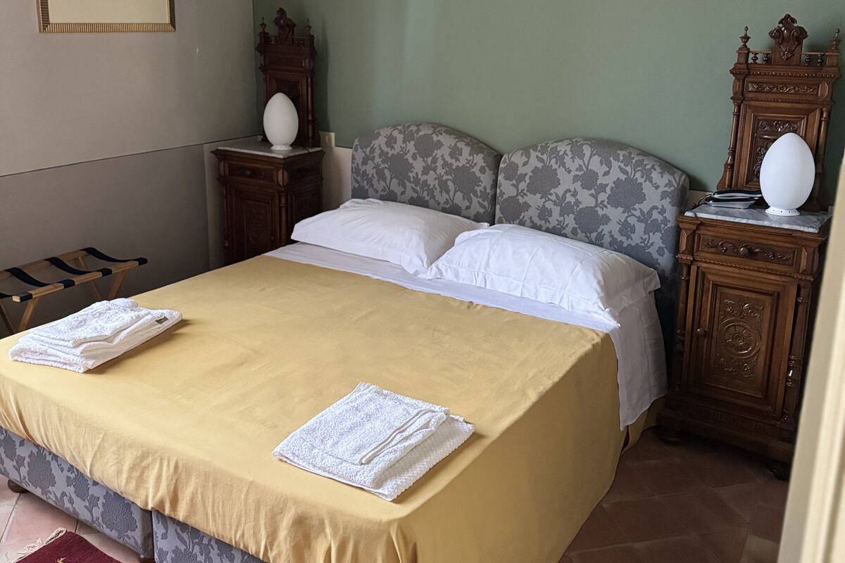 Un letto rifatto con copriletto giallo e cuscini bianchi, un comodino in legno con una lampada e un quadro appeso alla parete.