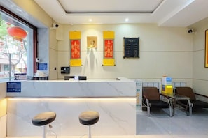 Zona de recepción de un hotel con mostrador de mármol, dos taburetes de bar, una mesa pequeña con sillas y paneles decorativos en la pared.