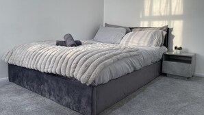 A modern bedroom with a grey upholstered bed, a fluffy white throw blanket, and a small bedside table with a plant. - 3 Bed House - Sleeps 5 - Garden - Parking - Wifi (Dartford)