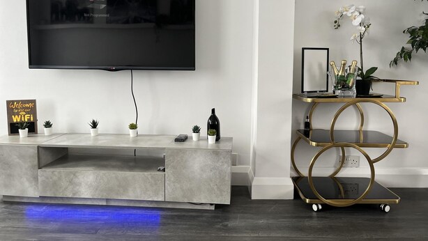 A modern living room with a flat-screen TV mounted on the wall, a grey entertainment unit with small potted plants, a gold-framed side table with a laptop and decorative items, and a blue-lit floor. - 3 Bed House - Sleeps 5 - Garden - Parking - Wifi (Dartford)