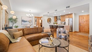 A modern living room with a brown leather sofa, a glass coffee table, and a dining area with wooden cabinets and a refrigerator. - Mountain Escape | Prime Location | Stunning Views (Canmore)