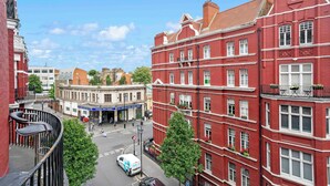 Ein rot gebackenes Gebäude mit zahlreichen Fenstern und Balkonen, eine Straße mit Autos und Fußgängern sowie ein klarer, blauer Himmel.
