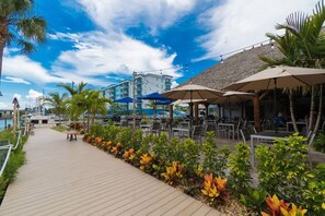 Un sendero de madera que conduce a un área de estar al aire libre con sombrillas, rodeado de plantas tropicales y vistas a un edificio con techo de paja.