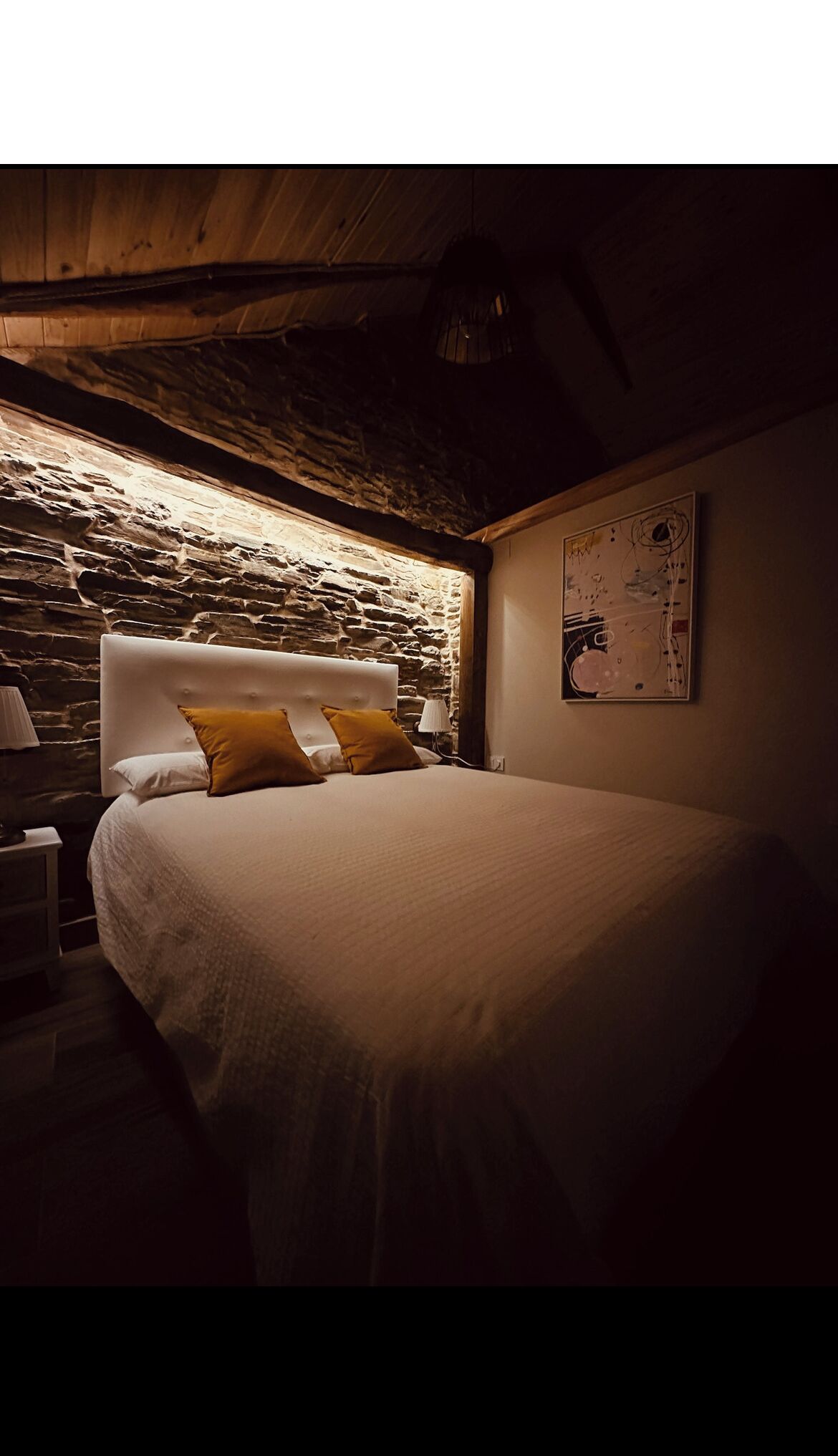 Une chambre avec un mur en pierre, un lit avec une literie blanche et deux oreillers, une table de chevet avec une lampe et un tableau accroché au mur.