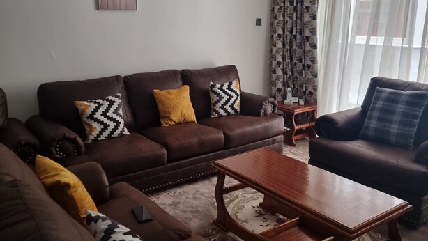 A living room with a brown sofa, a wooden coffee table, and a patterned rug. - Safari City Two Bedroom Apartment (Nairobi)