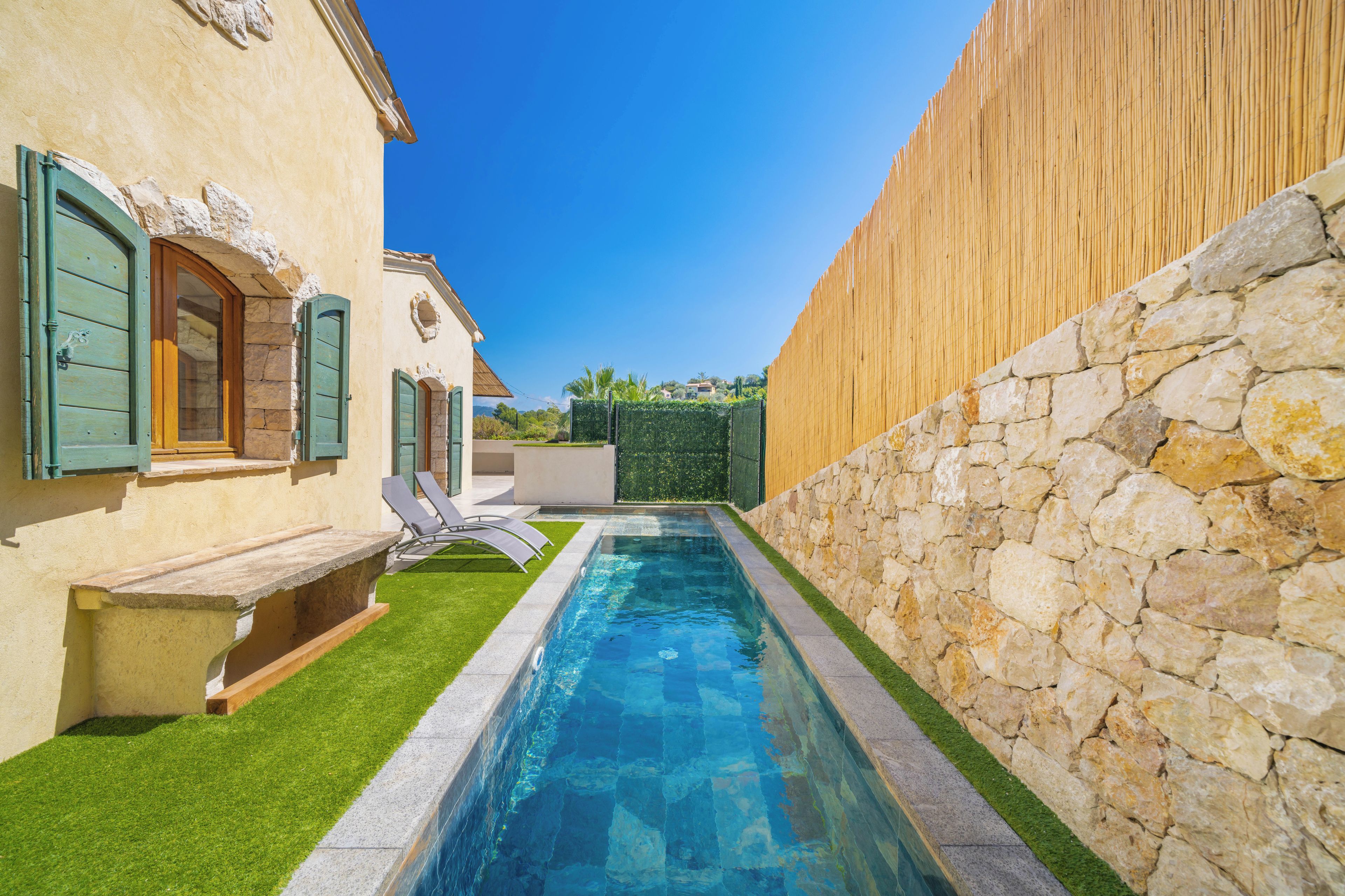 A swimming pool with a stone wall on one side and a building with a window and shutters on the other.