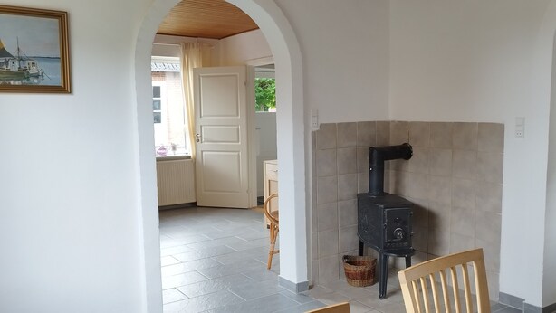 A room with a tiled floor, a wooden chair, a black stove, a white door, and a painting of a boat. - Beautiful town house with room for 5 people close to the center of Nykøbing Mors (Nykøbing Mors)