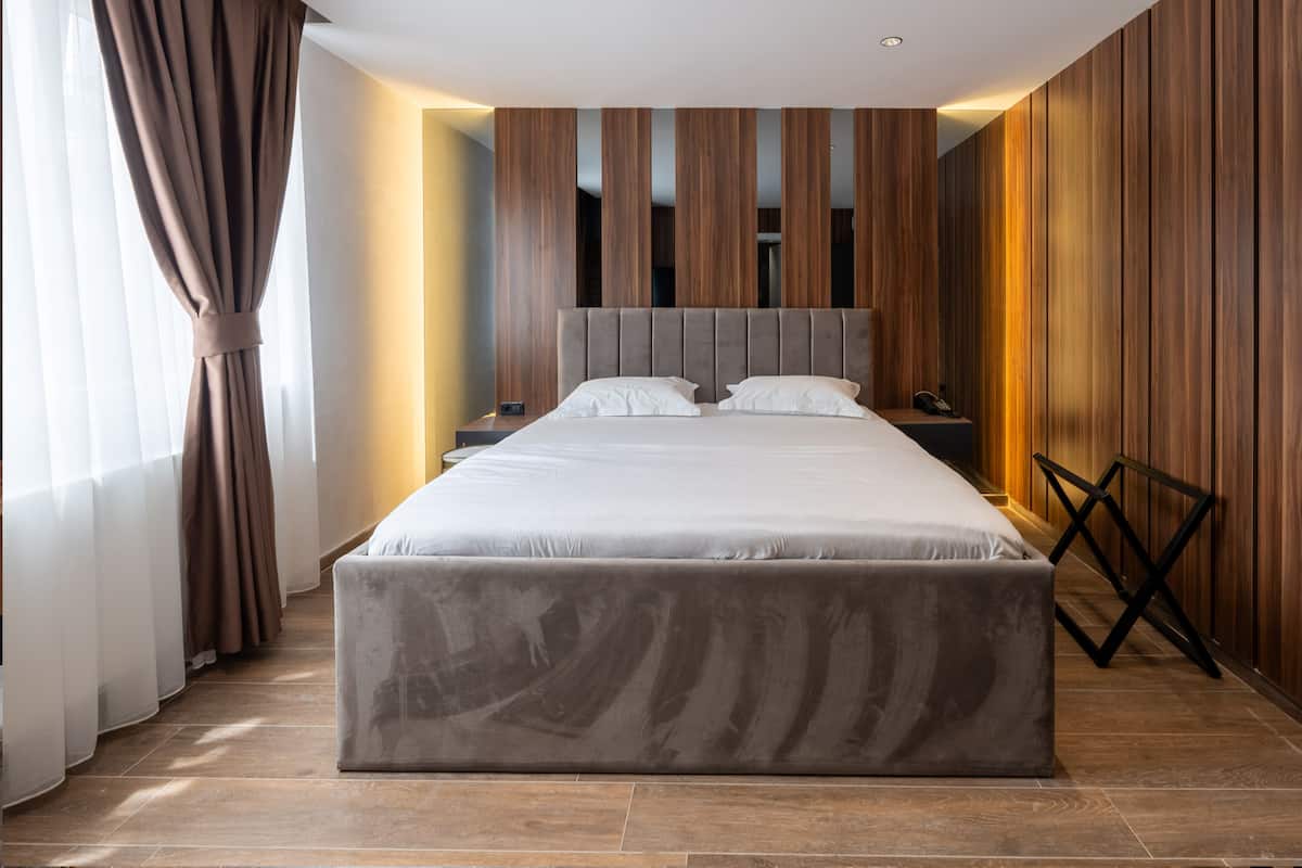 A modern hotel room with a large bed, wooden panel walls, a nightstand, and a cross-back chair.