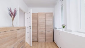 A modern room with a wooden floor, white walls, a wooden wardrobe, and a small plant on the windowsill. - Comfortable Apartment for 4 people | Close to Tauron Arena (Kraków)