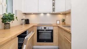 A compact kitchen with white cabinets, a built-in oven, a dishwasher, and a window. - Comfortable Apartment for 4 people | Close to Tauron Arena (Kraków)