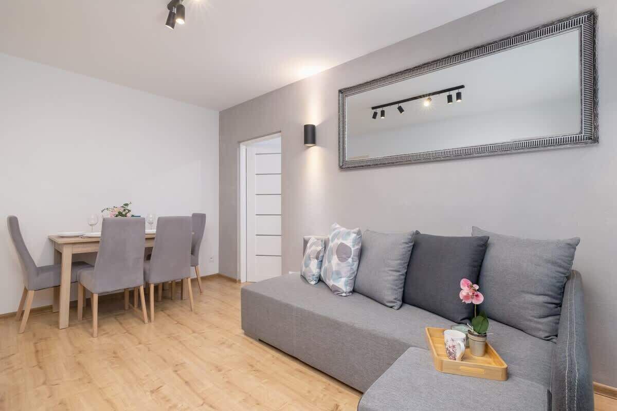 A modern living room with a grey sofa, a dining table with chairs, and a large mirror on the wall.