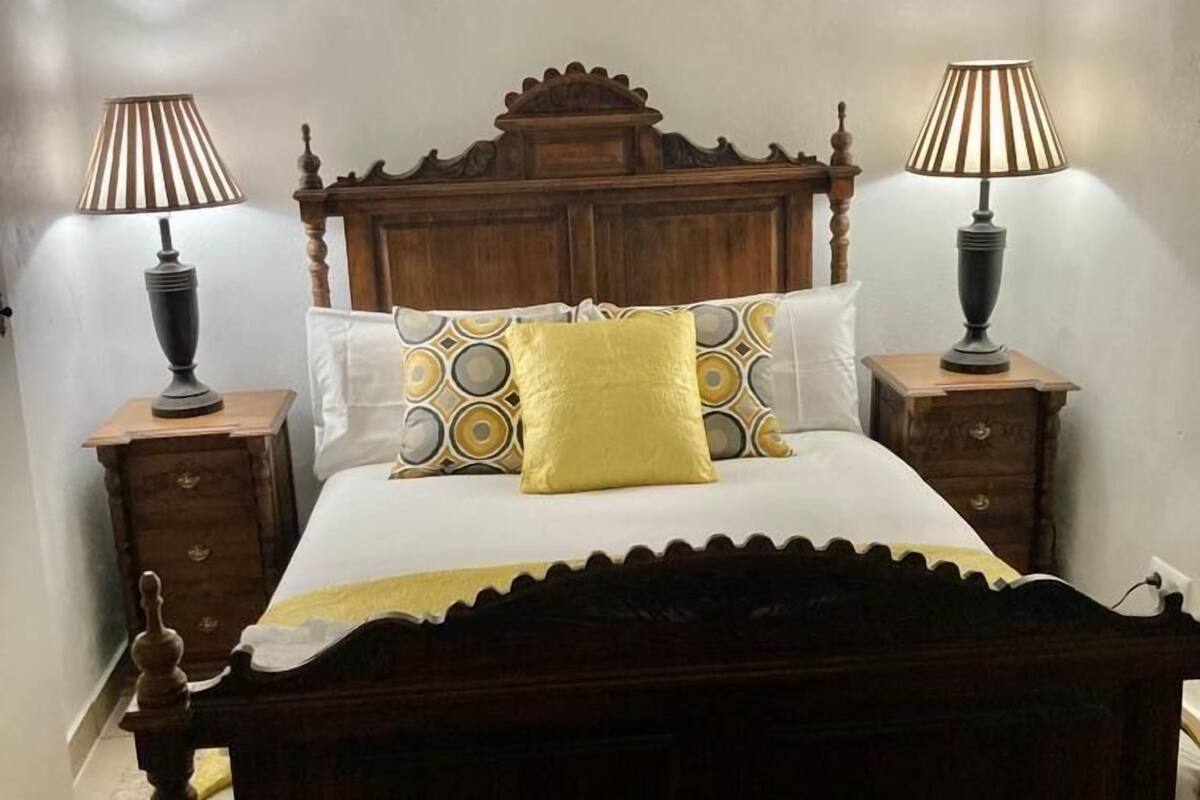 A wooden four-poster bed with a white and yellow patterned bedspread, flanked by two bedside tables with lamps, in a room with exposed wooden beams on the ceiling.