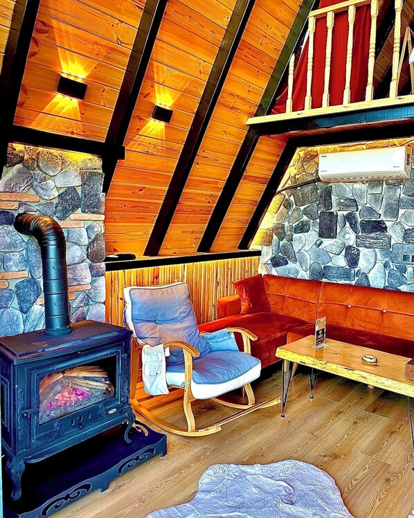 Una habitación acogedora con pared de piedra, techo de madera, chimenea, sillón mecedora, sofá rojo y una mesa.