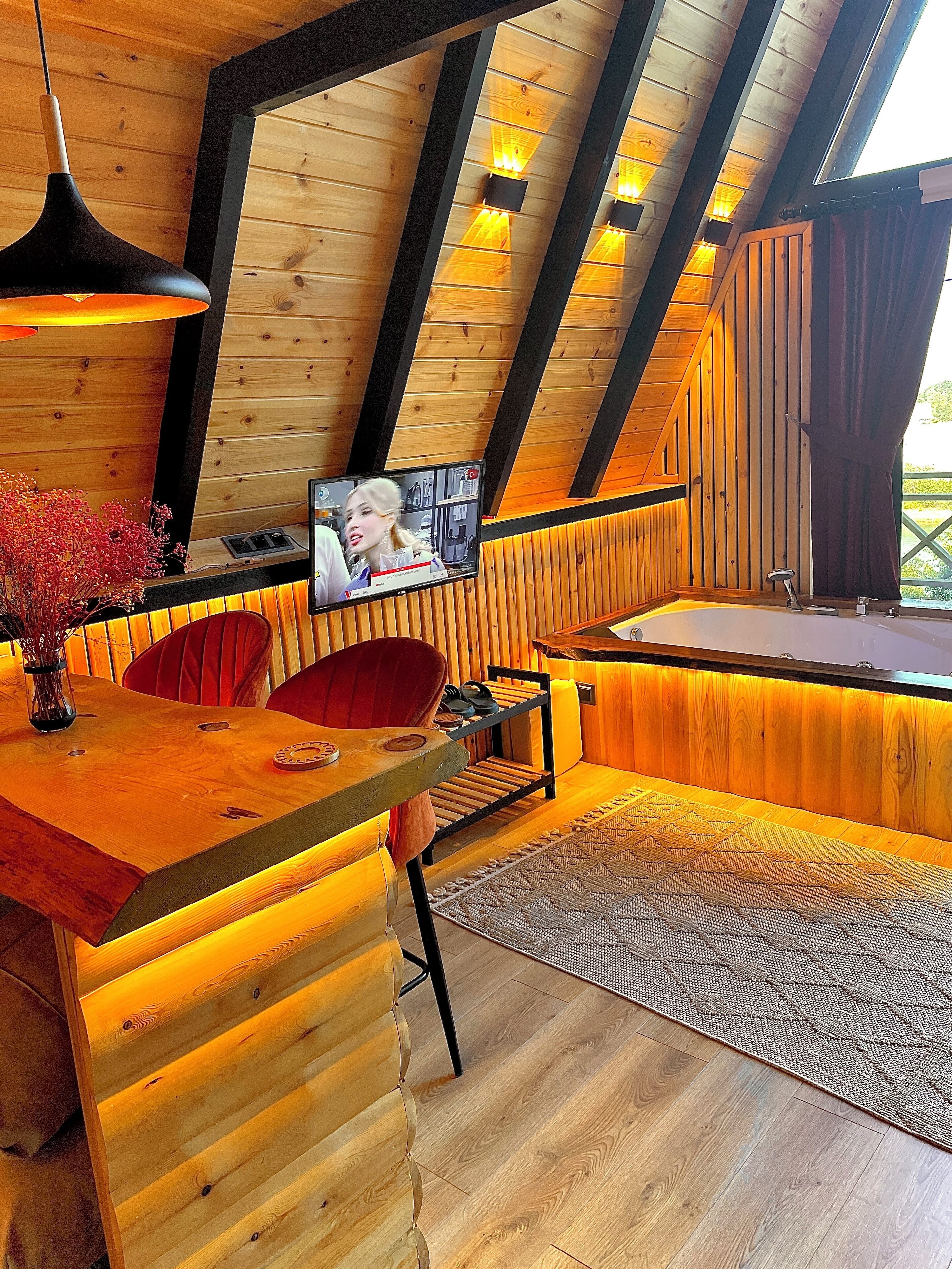 A cozy, wooden interior with a wooden desk, a red chair, a television, and a large window.