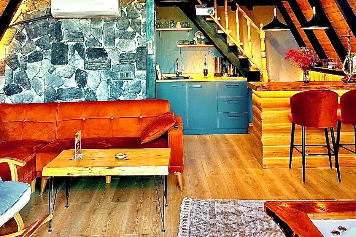 Un espace de vie de style « loft », avec un plafond en bois, un mur en pierre, un coin cuisine et un espace détente comprenant un canapé et des fauteuils.