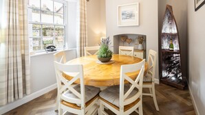 A dining area with a wooden table and white chairs, a potted plant, a wine rack, and a window with curtains. - Host Stay Arundel Cottage (Whitby)