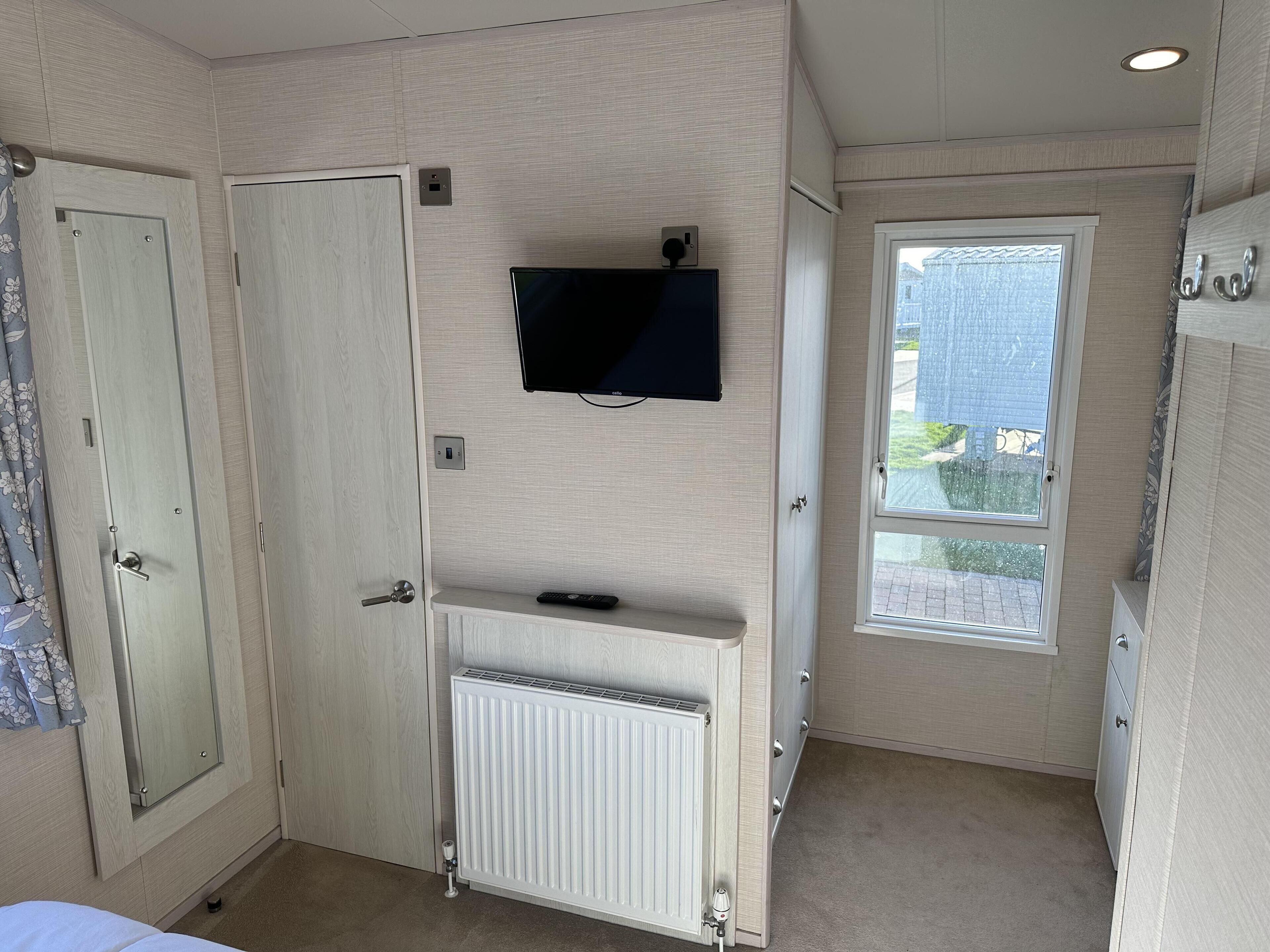 Quarto pequeno e bem iluminado, com uma cama, radiador, TV fixada na parede e janela com vista para o exterior.