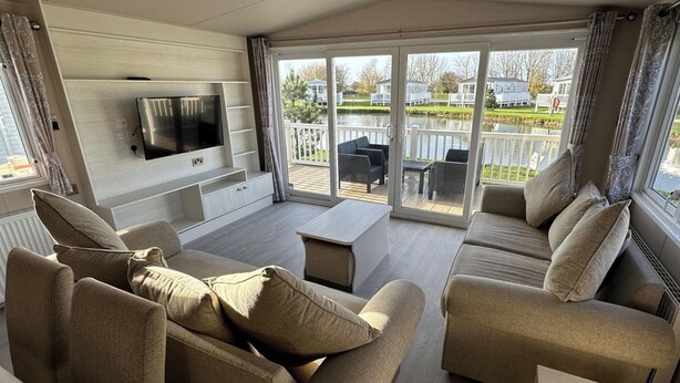 A modern living room with a flat-screen TV, beige sofas, a coffee table, and a sliding glass door leading to an outdoor patio with a view of houses and a lake. - Mablethorpe Lakeside Caravan With Decking & Wi-fi! (Mablethorpe)