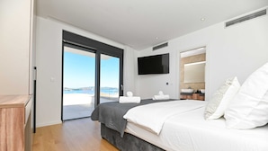 A modern bedroom with a large bed, a flat-screen TV, and a view of the sea through a sliding door. - Villa Oasis (Trogir)