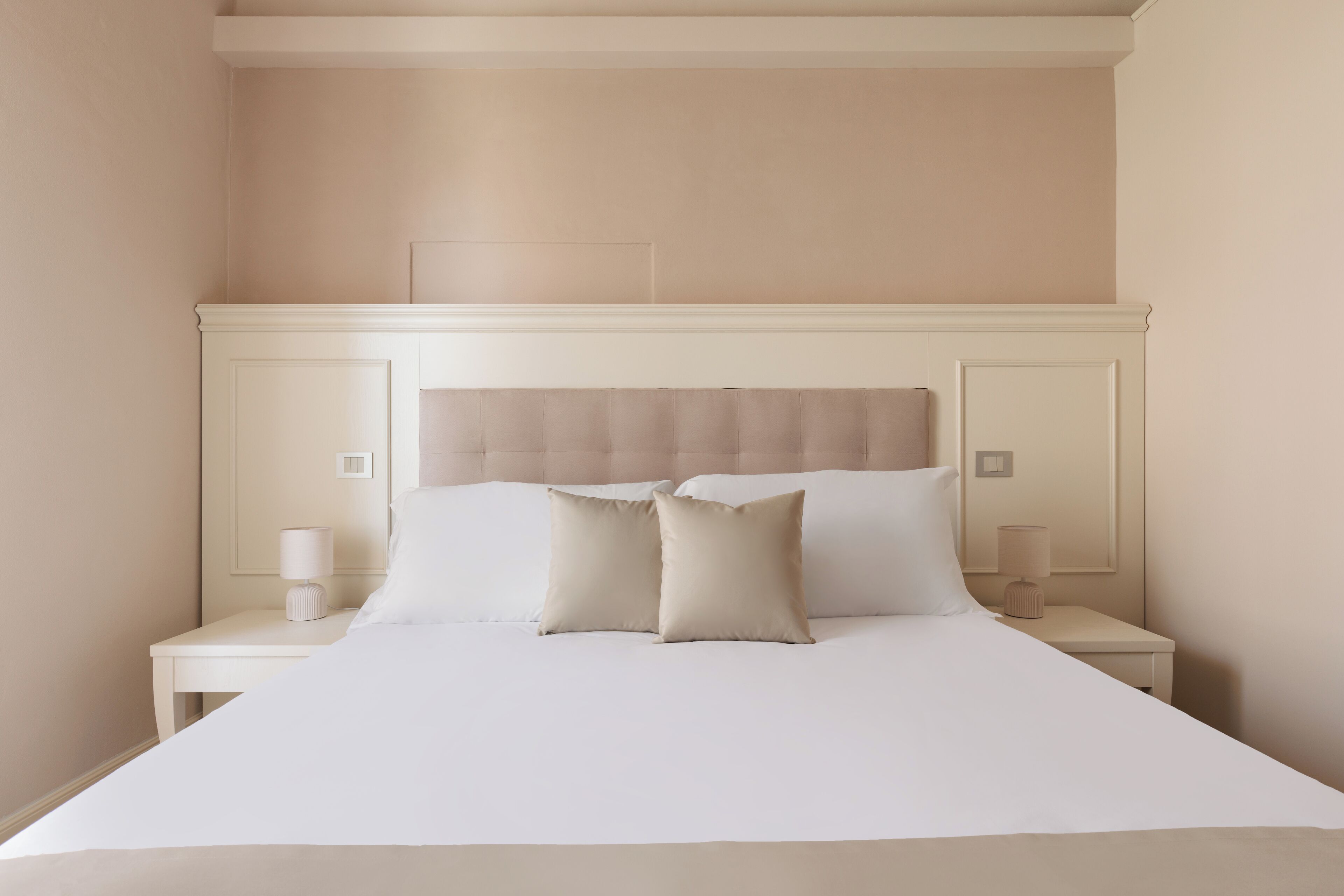 Una cama bien tendida con sábanas blancas y dos almohadas, flanqueada por dos mesitas de noche con lámparas, en una habitación de diseño sencillo y limpio.