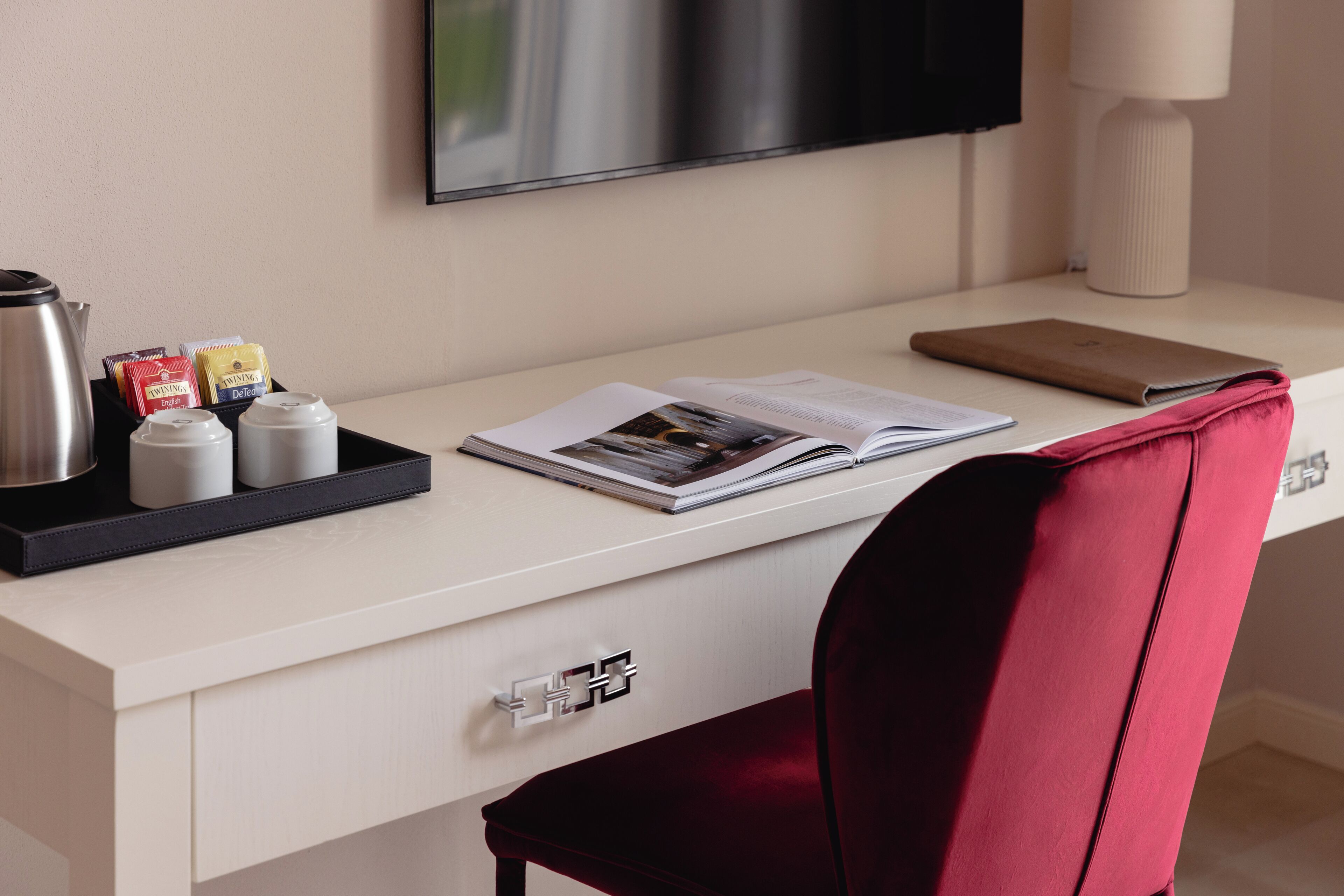 A modern hotel room with a white desk, a red upholstered chair, a flat-screen TV mounted on the wall, and a bedside table with a lamp.