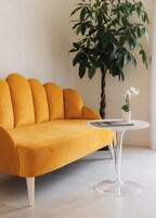 Un sofá de color mostaza con patas blancas, una mesa redonda blanca con tapa de cristal transparente y una planta en maceta con flores blancas.