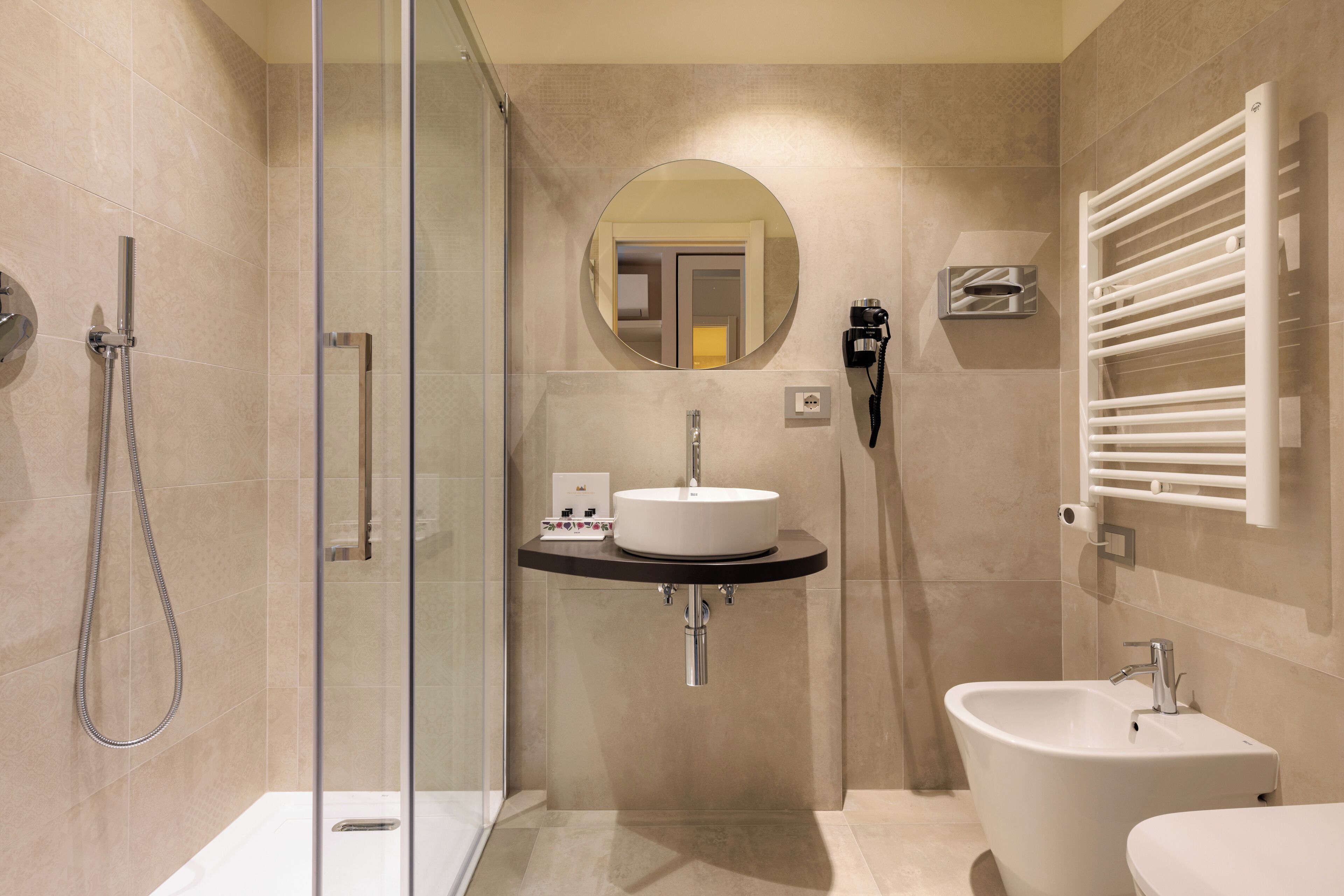 A modern bathroom with a glass shower enclosure, a round mirror, a sink with a towel rack, and a bidet.