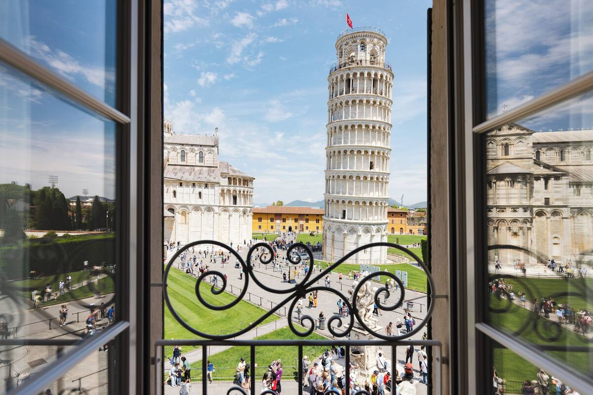 A view of the Leaning Tower of Pisa through a window with a decorative iron grille.