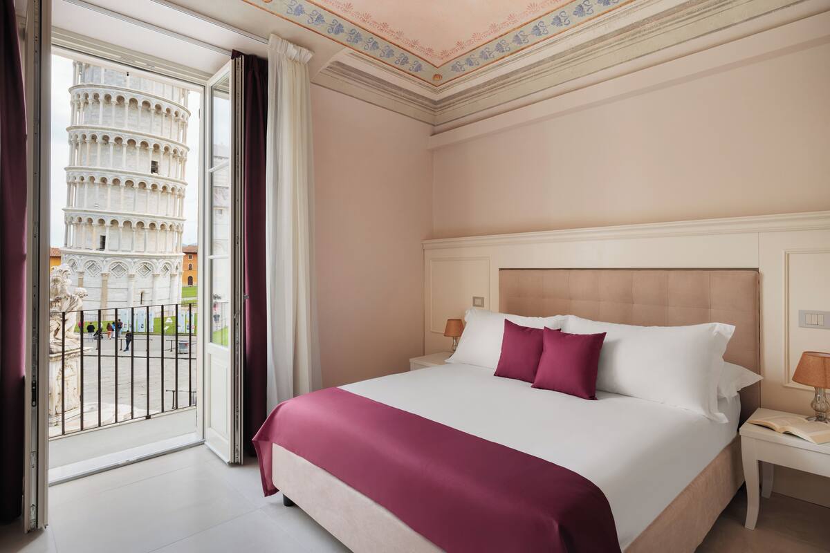 A hotel room with a large bed, a nightstand, a window with a view of the Leaning Tower of Pisa, and a balcony with a railing.