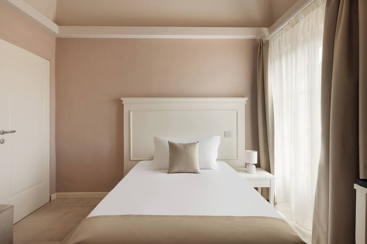 A hotel room with a large bed, a bedside table with a lamp, and a view of the outside through sheer curtains.