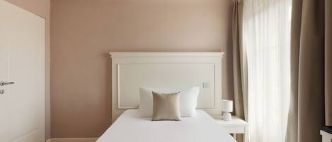 A hotel room with a large bed, a bedside table with a lamp, and a view of the outside through sheer curtains.