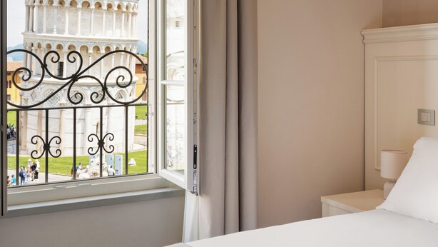 A hotel room with a bed, a nightstand, a lamp, and a window with a view of the Leaning Tower of Pisa. - Prato dei Miracoli Residenza d'Epoca (Pisa)