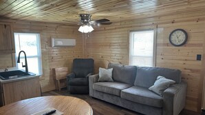 Een gezellig cabin interieur met een houten plafond, een plafondventilator, een grijze bank, een houten eettafel, een keuken met een spoelbak en raam, en een wandklok.