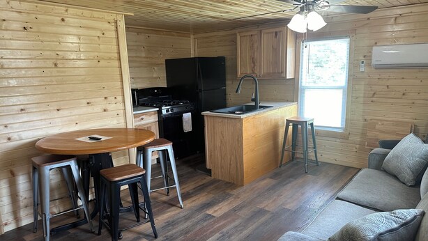 Een compacte keuken met een fornuis, spoelbak en koelkast, een houten eethoek met krukken, en een grijze bank in een interieur in chaletstijl.
