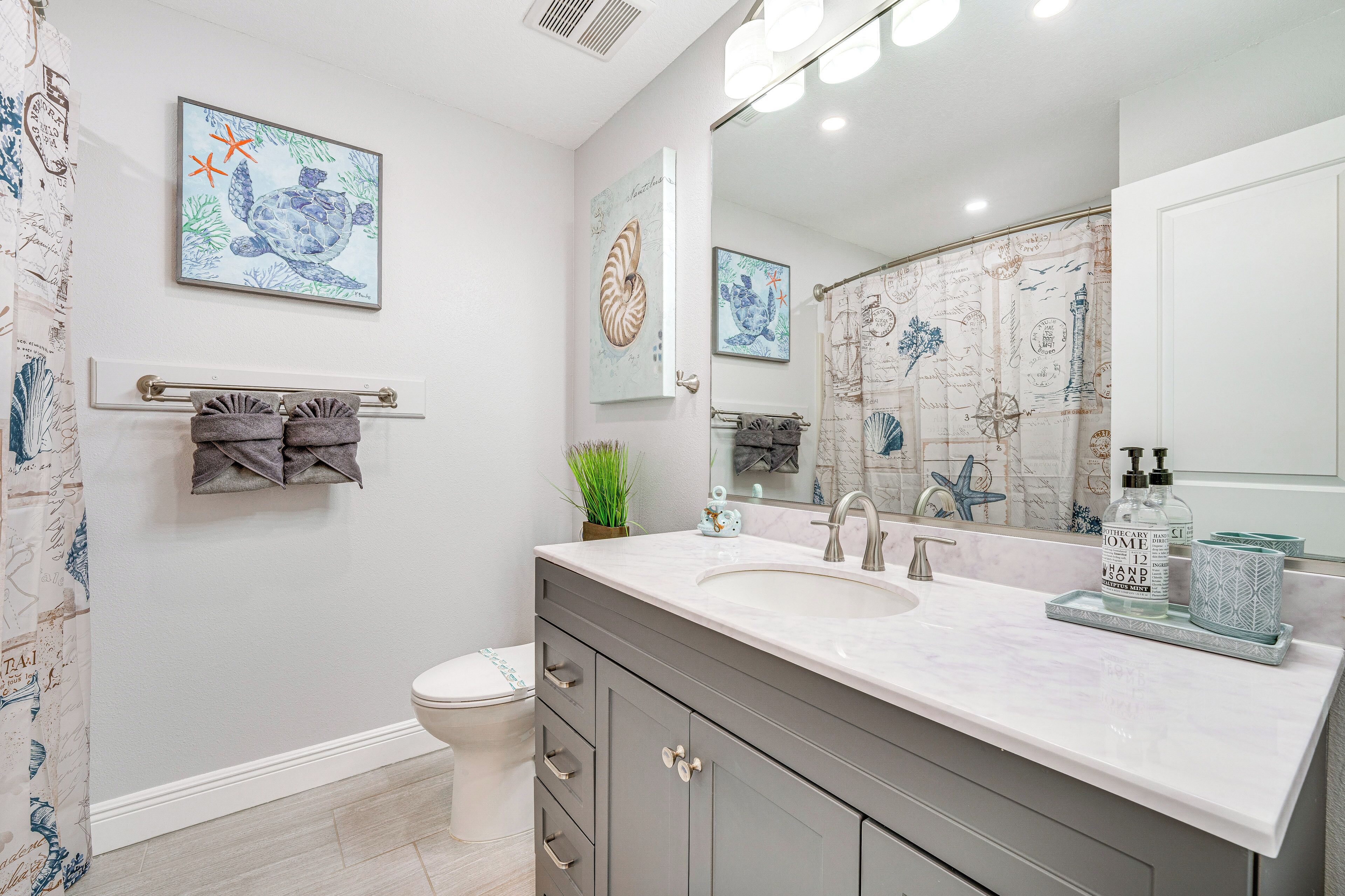 Une salle de bain avec un lavabo blanc, des meubles gris et un grand miroir. On y trouve deux tableaux encadrés accrochés au mur, un porte-serviettes avec des serviettes et une plante en pot.