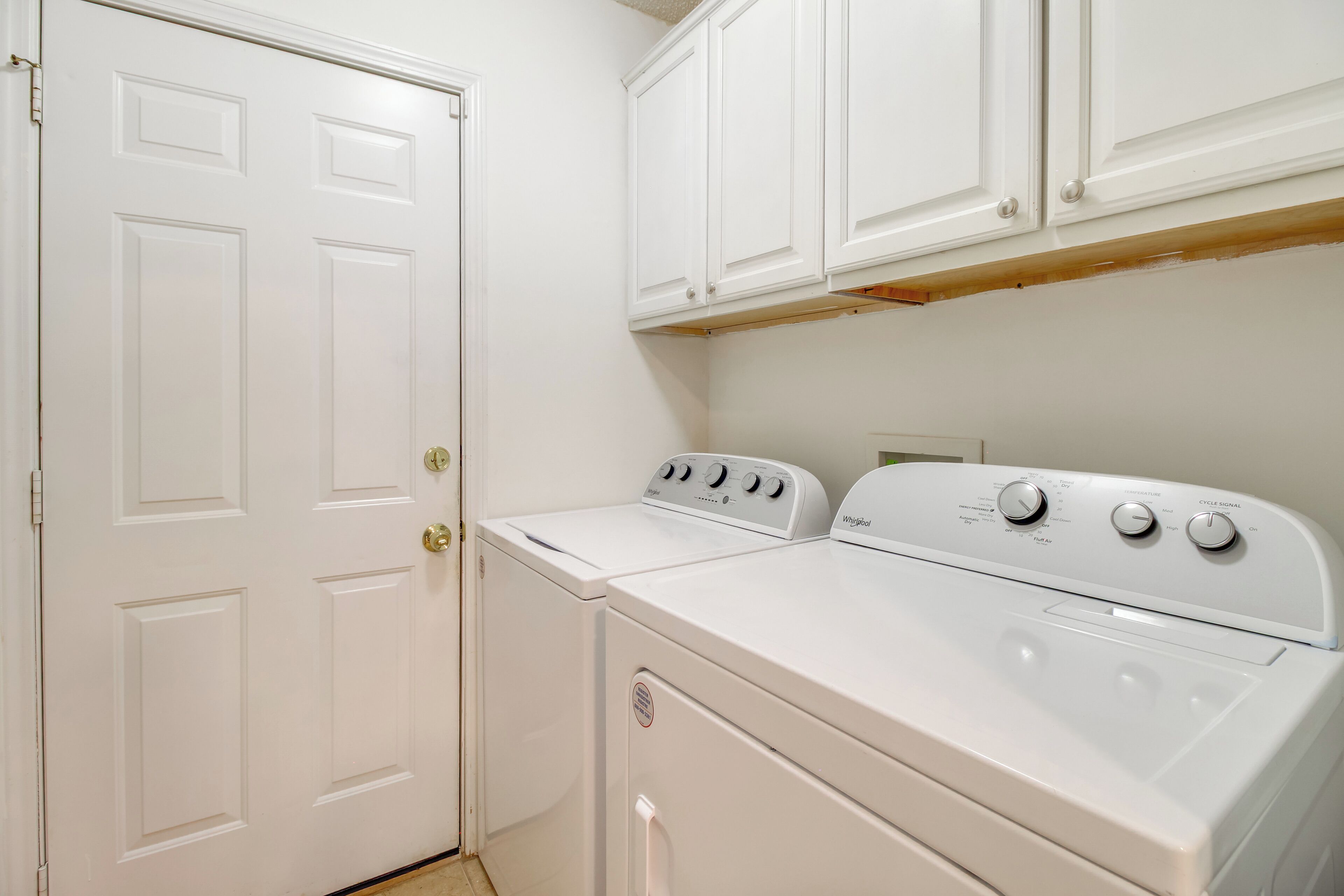 A laundry room with a front-loading washer and dryer, white cabinets above, and a door to the left.