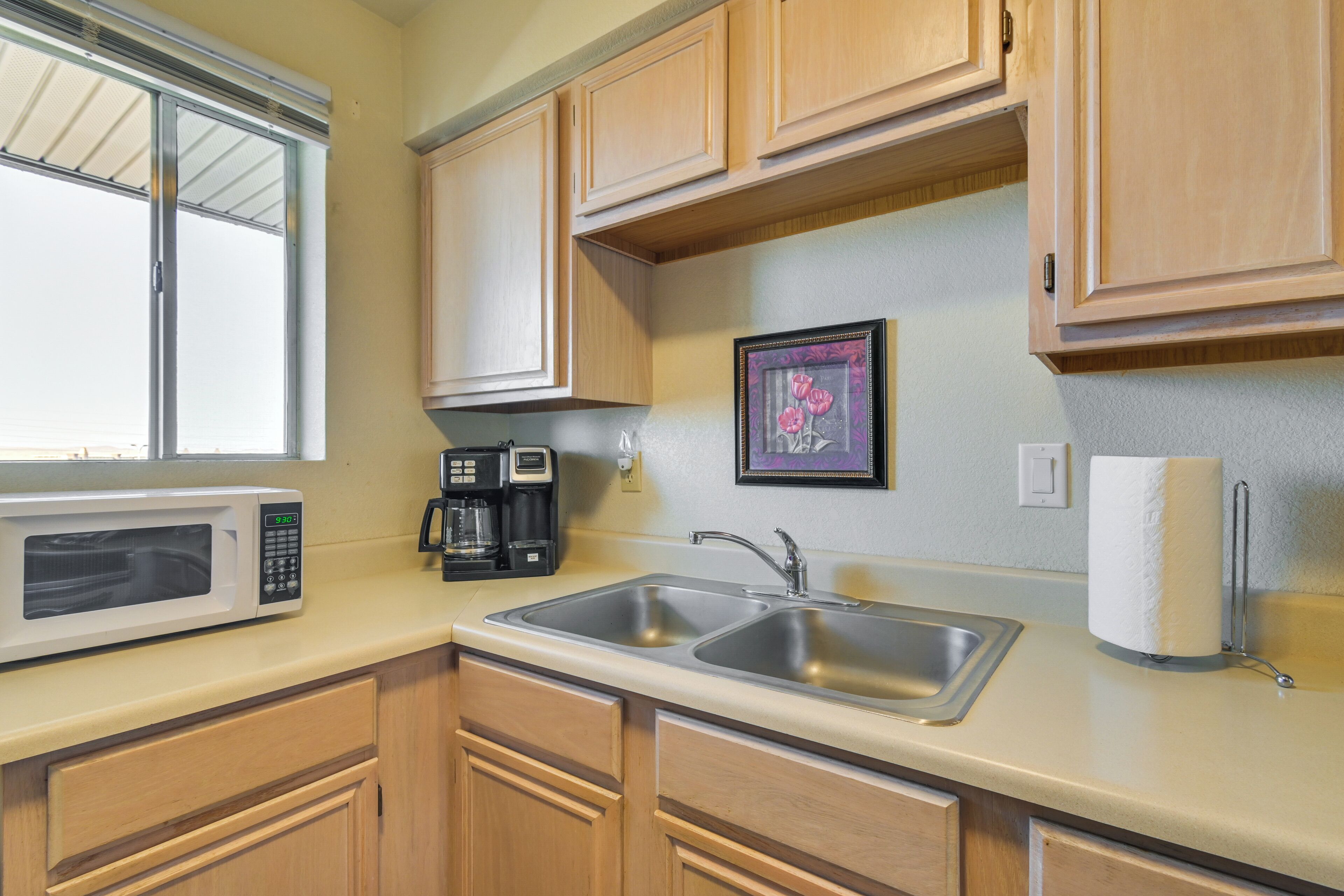 A kitchen with wooden cabinets, a stainless steel sink, a microwave, a coffee maker, and a framed picture on the wall.