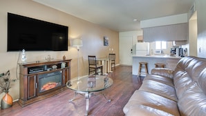 A living room with a fireplace, a glass coffee table, a leather sofa, a flat-screen TV, and a kitchen area with a microwave.